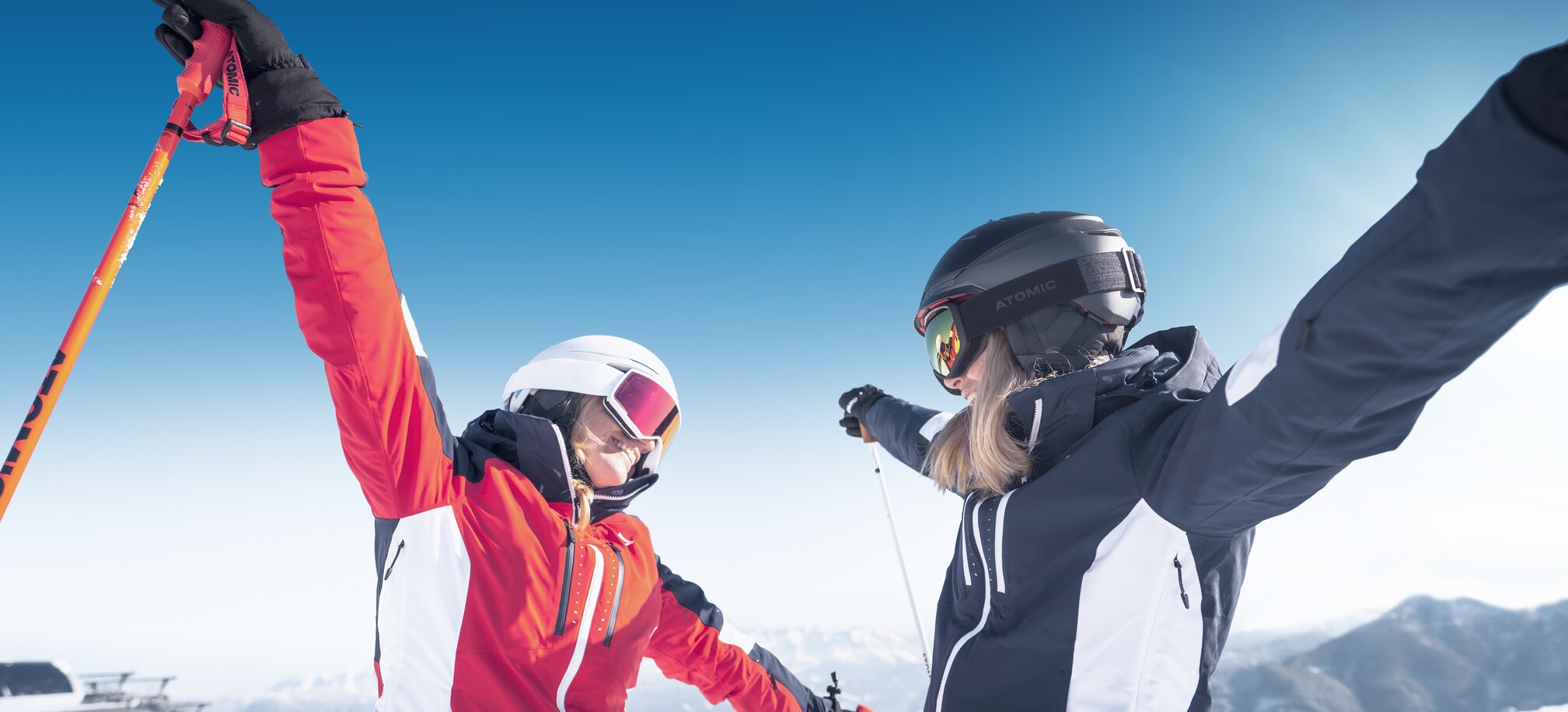 Two female skiers face each other with arms wide open, smiling in the snow and sunshine