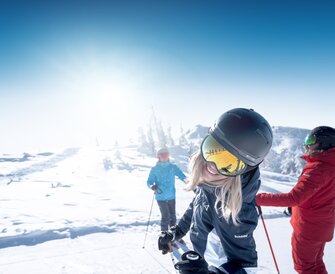 Woman with helmet and ski goggles smiles at camera, two skiers in snow behind her
