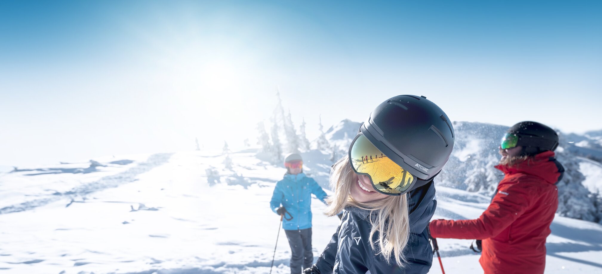 Woman with helmet and ski goggles smiles at camera, two skiers in snow behind her