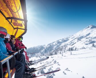 Five skiers sit on a chairlift with orange cover above a snowy mountain landscape