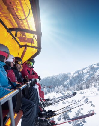 Five skiers sit on a chairlift with orange cover above a snowy mountain landscape