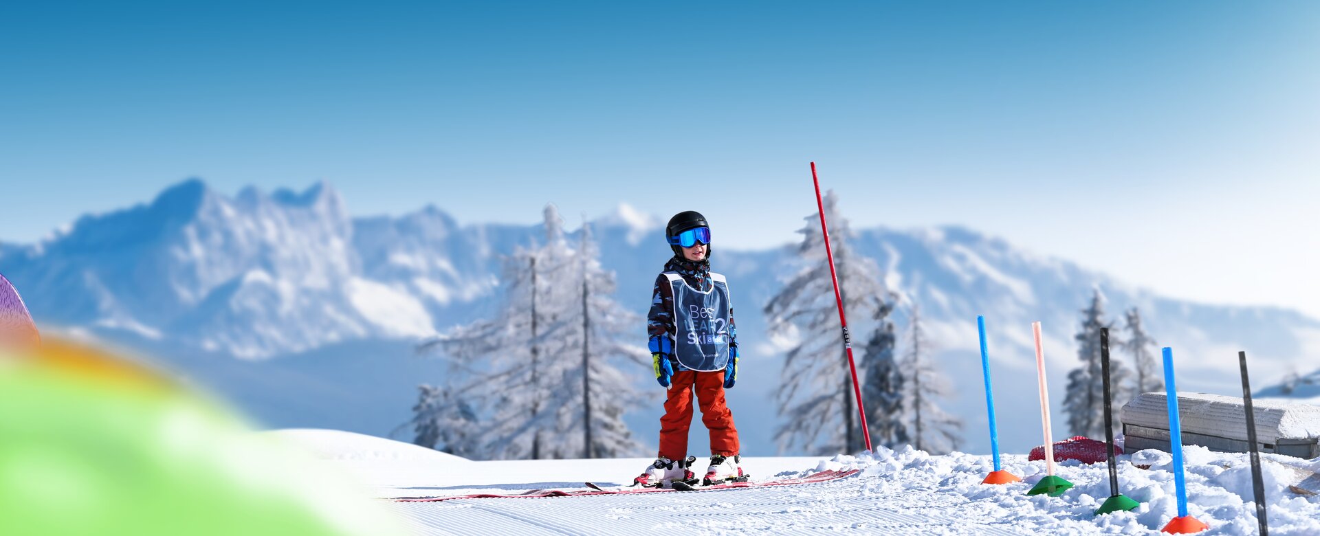 Child wearing helmet and vest stands on sunny training slope with skis in front of snowy mountains