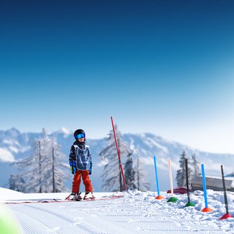 Child wearing helmet and vest stands on sunny training slope with skis in front of snowy mountains
