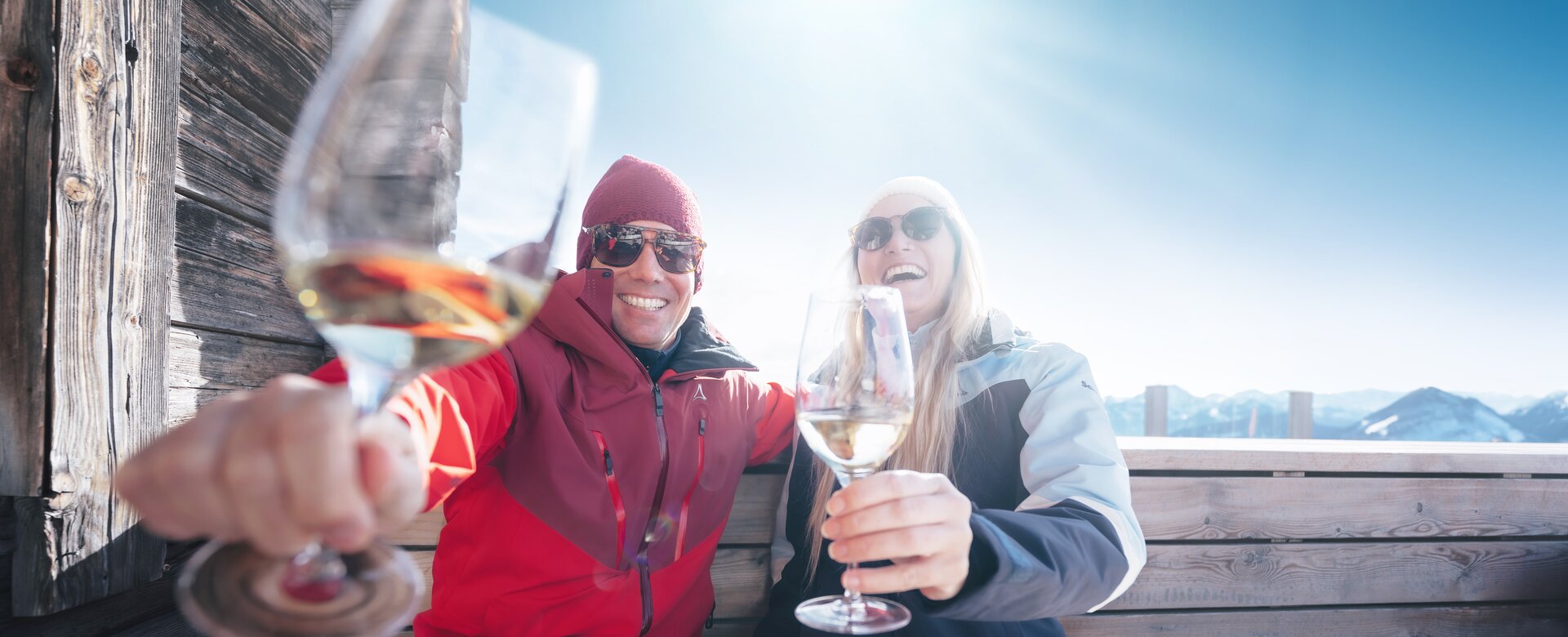 Two skiers toast with white wine on bench at hut in Ski amadé, sun and mountains in background
