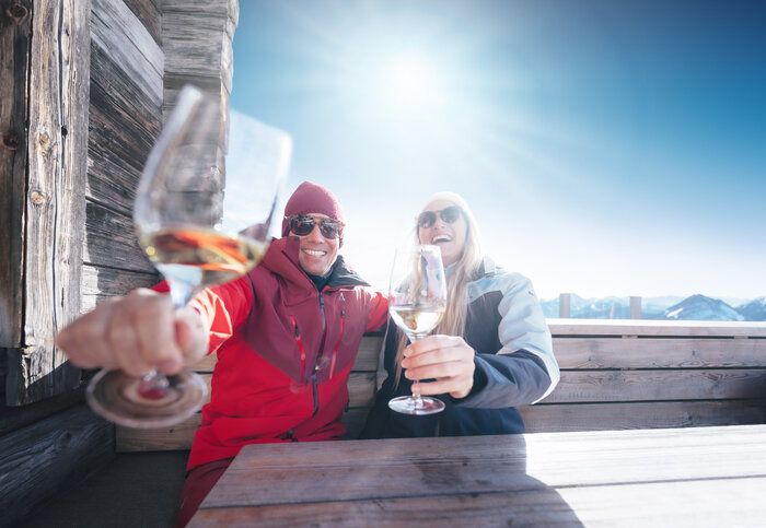 Two skiers toast with white wine on bench at hut in Ski amadé, sun and mountains in background
