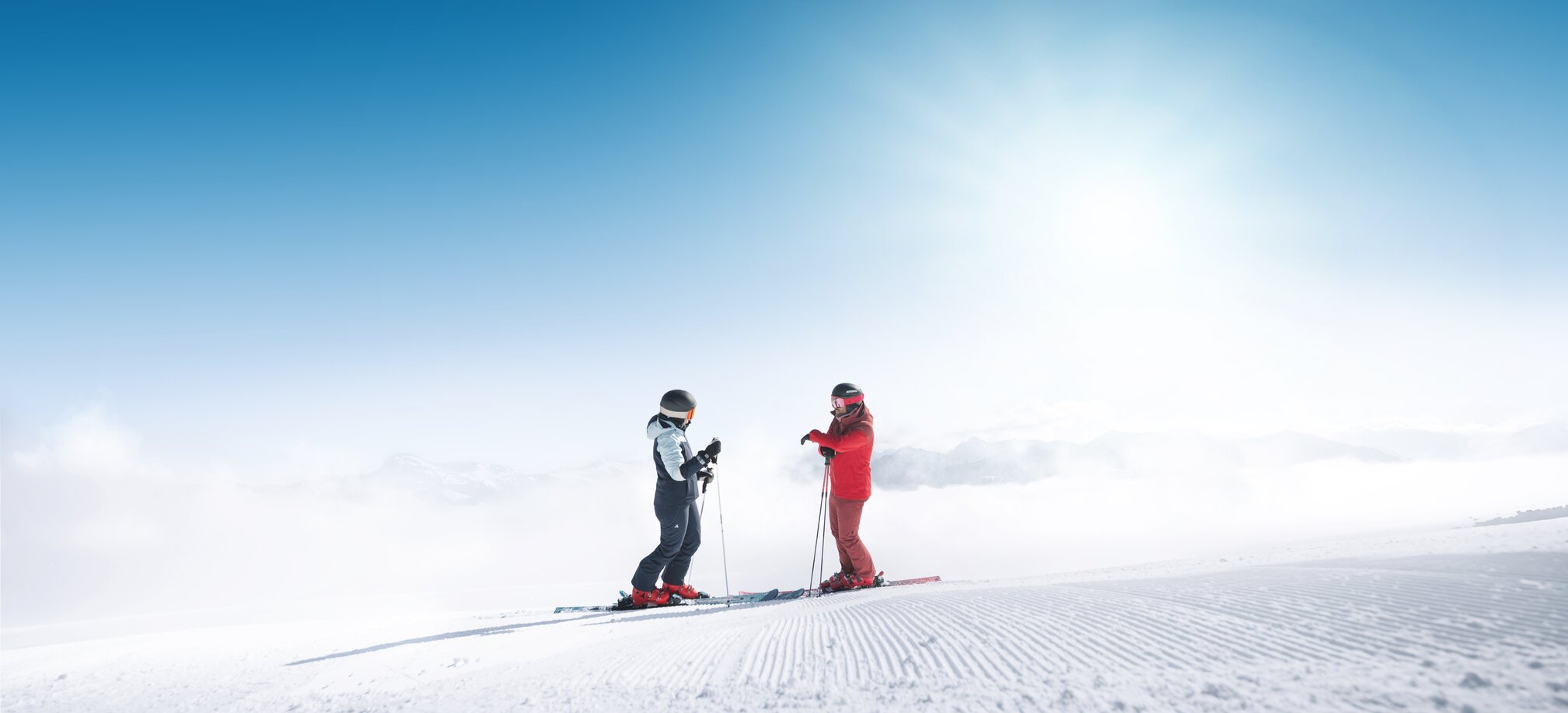 Two skiers stand on freshly groomed slope in Ski amadé overlooking a foggy valley in bright sunlight
