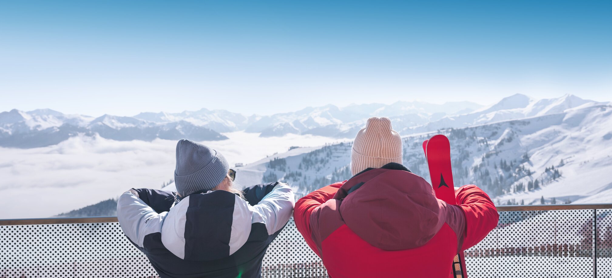 Two people in winter clothes stand on terrace overlooking snowy mountains with skis in the foreground
