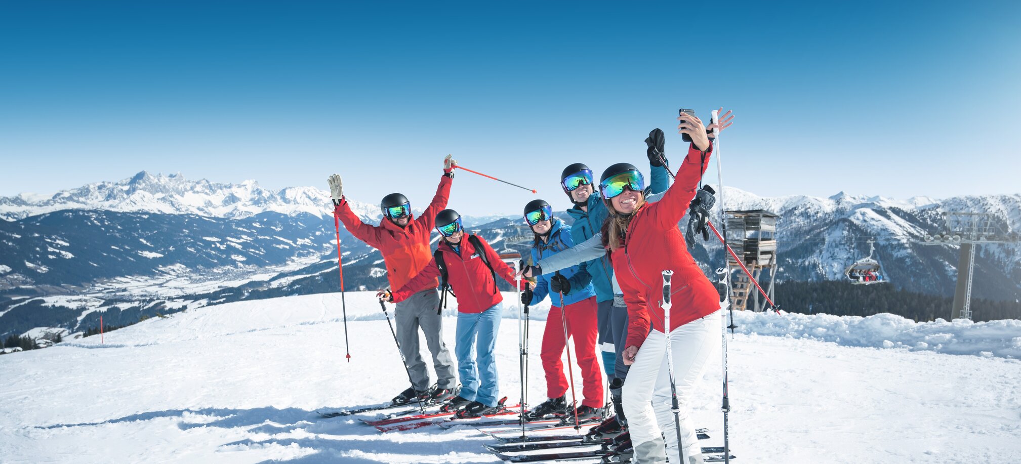 Sechs Skifahrer machen bei Sonnenschein auf der Piste ein fröhliches Gruppen-Selfie