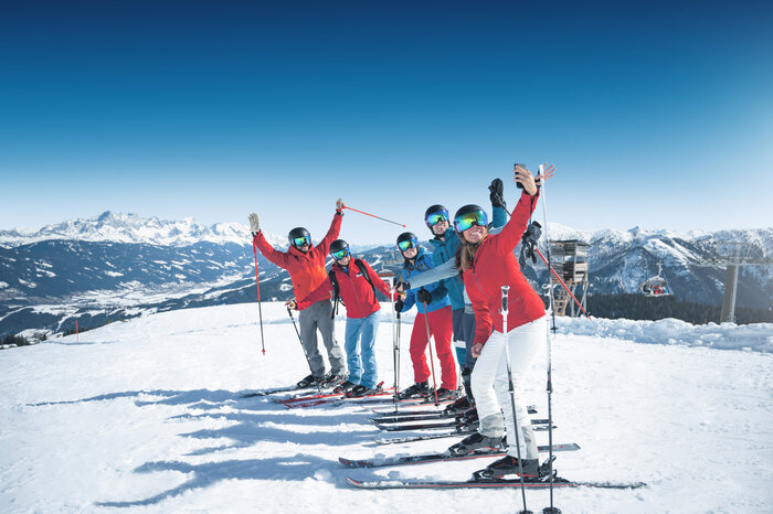 Six skiers take a cheerful group selfie on the slope in sunny weather with mountains behind