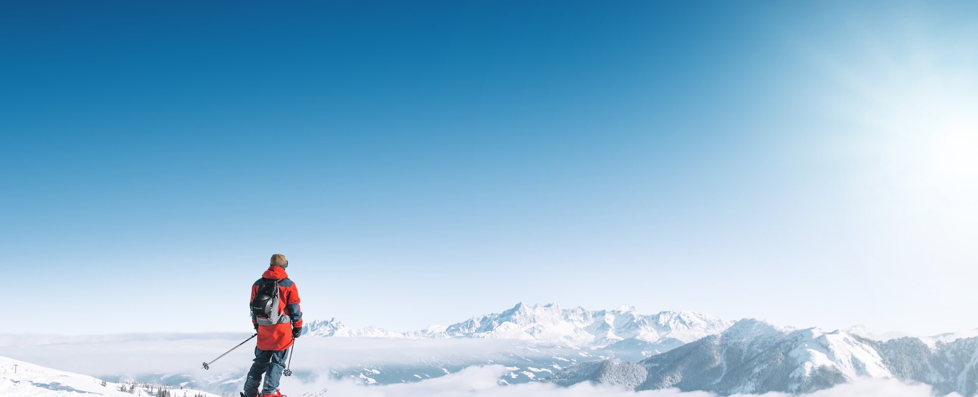 A skier looks out over snowy mountains and a vast sea of clouds in bright winter sun