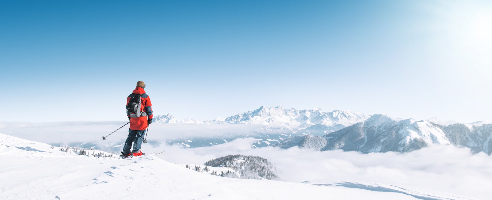 A skier looks out over snowy mountains and a vast sea of clouds in bright winter sun
