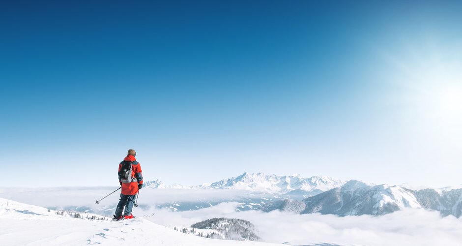 A skier looks out over snowy mountains and a vast sea of clouds in bright winter sun