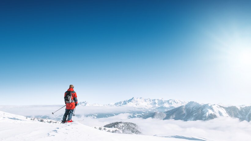 A skier looks out over snowy mountains and a vast sea of clouds in bright winter sun