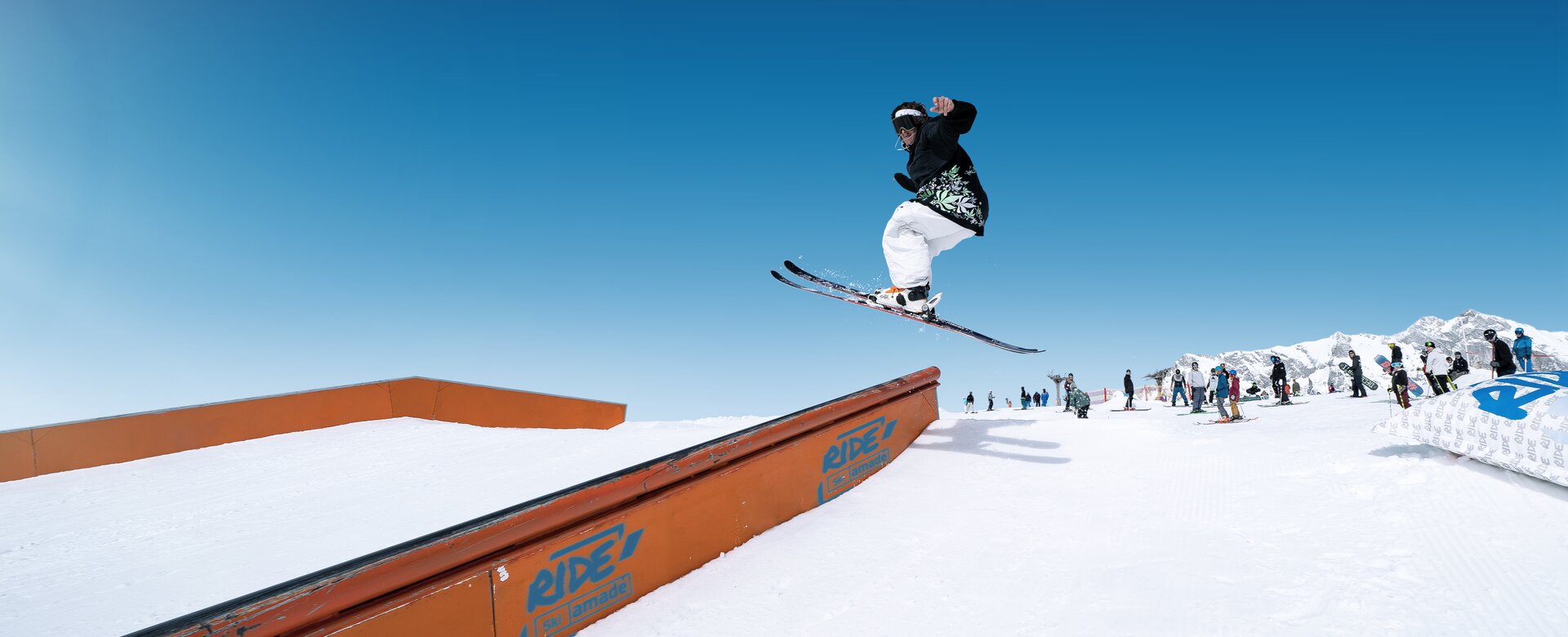 Skier jumps over rail element in snow park, other winter sports fans watching in the background