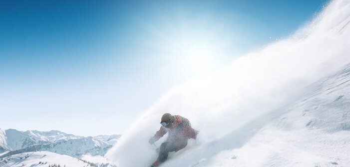 Snowboarder rides down steep slope in deep powder snow, spray clouds in the sunlight