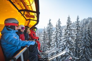 Four skiers wearing helmets and goggles sit on a chairlift with an orange cover above snow-covered fir trees. | © Ski amadé