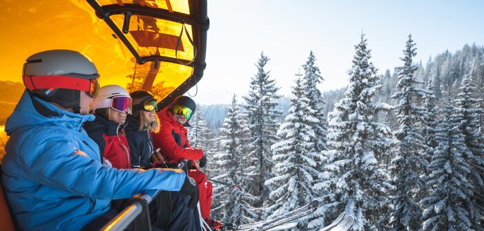 Four skiers wearing helmets and goggles sit on a chairlift with an orange cover above snow-covered fir trees. | © Ski amadé