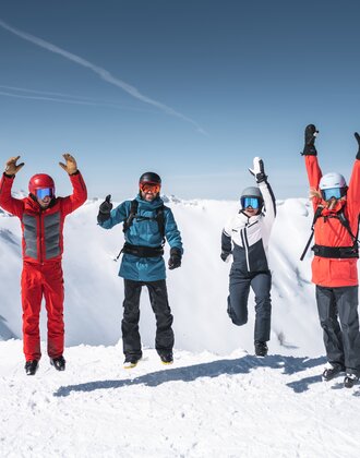 Vier Skifahrer springen gleichzeitig in die Luft, Schneeberge und Sonne im Hintergrund