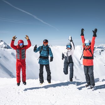 Four skiers jump in the air together with snowy mountain peaks and sunlight in the background