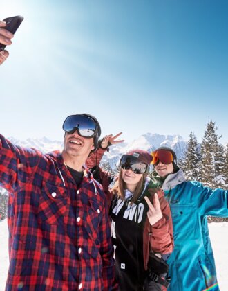 Drei jugendliche Skifahrer posieren für ein Selfie vor einer schneebedeckten Berglandschaft. | © Ski amadé
