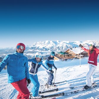 Four skiers laugh on the slope with a mountain hut in the background. | © Ski amadé