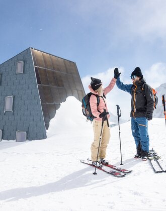 Two ski tourers high five in front of a modern hut in a snowy high mountain landscape  | © Harald Steiner