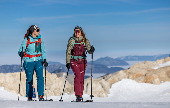 Two women hiking with ski touring gear and poles through a sunny snowy mountain area | © Harald Steiner