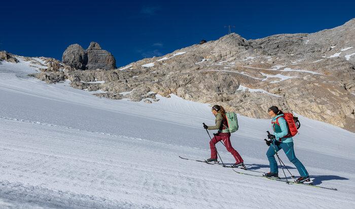 Two women ascend a snowy slope on touring skis below rocky mountain cliffs | © Harald Steiner