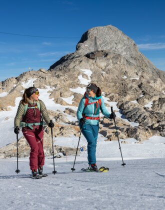 Two women ski touring in sunny weather with rocky mountain peak in the background | © H. Steiner