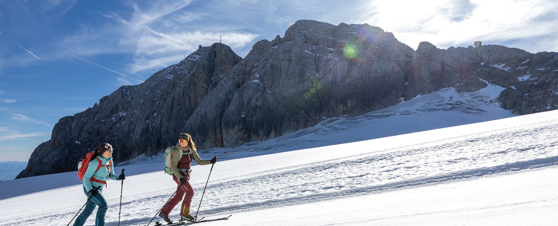 Two women ascend on touring skis in sunshine before a steep rocky mountain wall | © Harald Steiner
