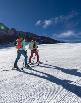 Two women ski touring on groomed piste in sun with long shadows and mountain backdrop | © Harald Steiner