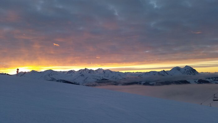 Verschneite Berghänge mit orangefarbenem Sonnenaufgang und Wolken über Mittelstation