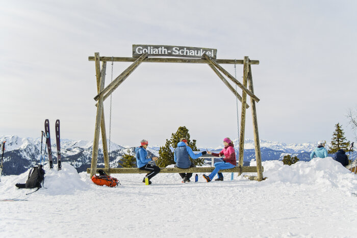 Ski tourers sit on large wooden swing with view of snowy alpine mountains