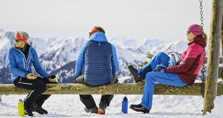 Three ski tourers sit with thermoses on wooden swing facing snowy mountain range