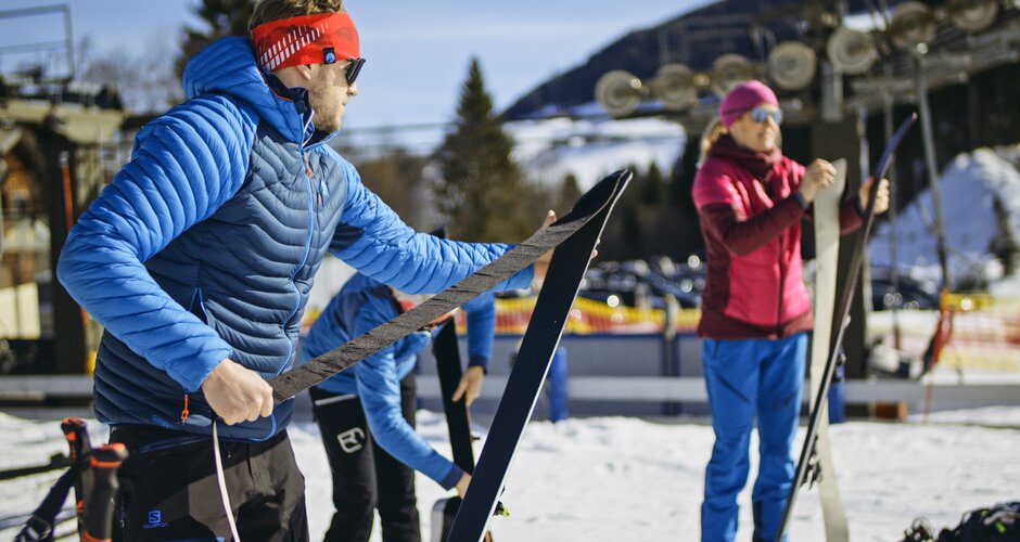 Two ski tourers attach climbing skins to their skis in sunshine near the slope