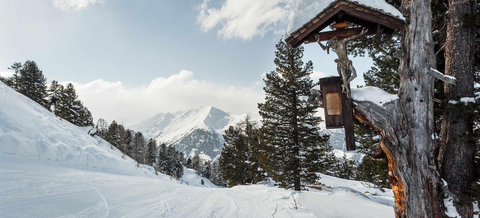 präparierter Weg mit einem Kreuz auf der Seite und Bäumen inmitten einer Schneelandschaft und Bergen. | © Georg Roske