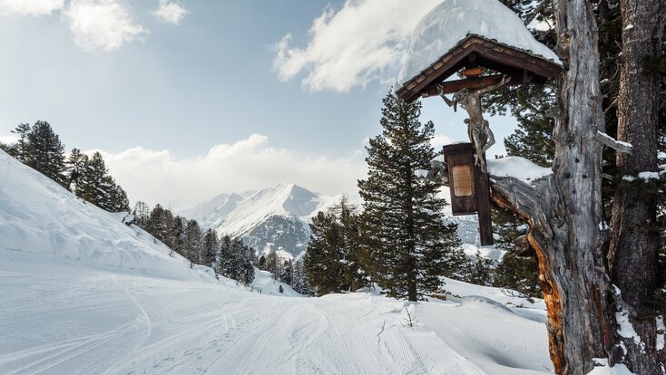 groomed path with a cross on the right and lined with trees amidst a snowy landscape and mountains. | © Georg Roske