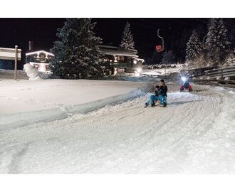 Family sledding at night on illuminated winter toboggan run with headlamps in Gastein, snowy hut in background | © GTG_Marktl Photography