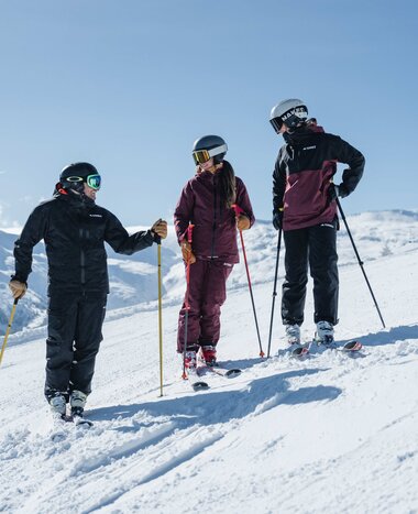 Three people in ski gear are standing on a slope talking with smiles | © Gasteinertal Tourismus GmbH, Christoph Oberschneider