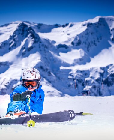 Child in blue ski gear sitting and laughing in snow with steep peaks in Gastein | © Max Steinbauer