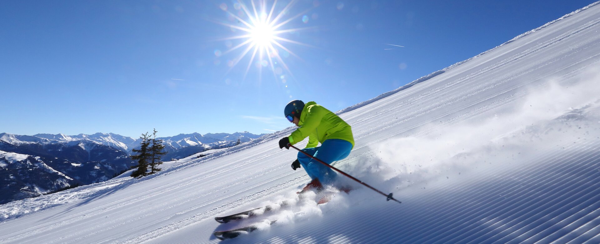 Skier skiing down a freshly groomed slope kicking up snow dust behind him and a snow-covered mountain panorama in the background | © Tourismusverband Großarltal