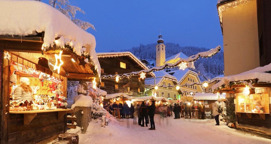 Lit advent stalls at Salzburg Mountain Advent in Grossarl with visitors, church and snow covered houses | © Tourismusverband Großarltal