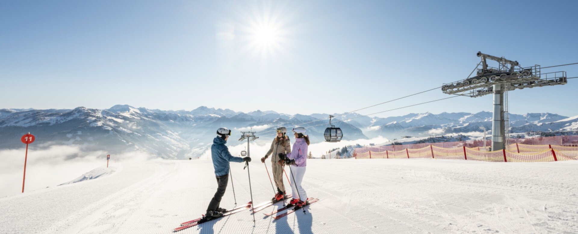 Three skiers stand on a freshly groomed slope in Grossarltal near a gondola lift with snowy Alps and bright sun | © Lorenz Masser