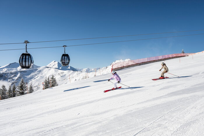 Two skiers descend a groomed slope in Grossarltal beneath a gondola lift with snowy mountains and blue sky | © Lorenz Masser