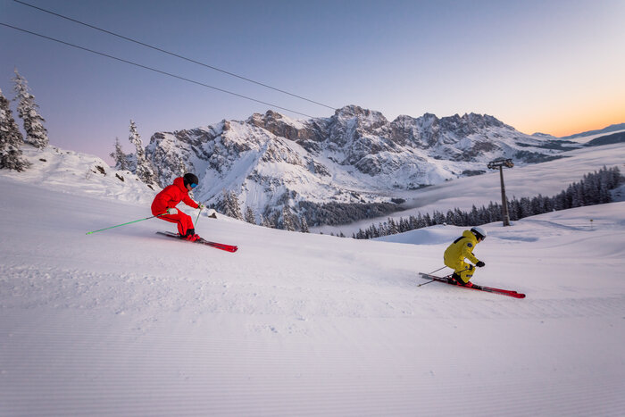 A skier in a yellow ski suit and one in red ski down the slope and the sun is just rising in the background | © Hochkönig Tourismus