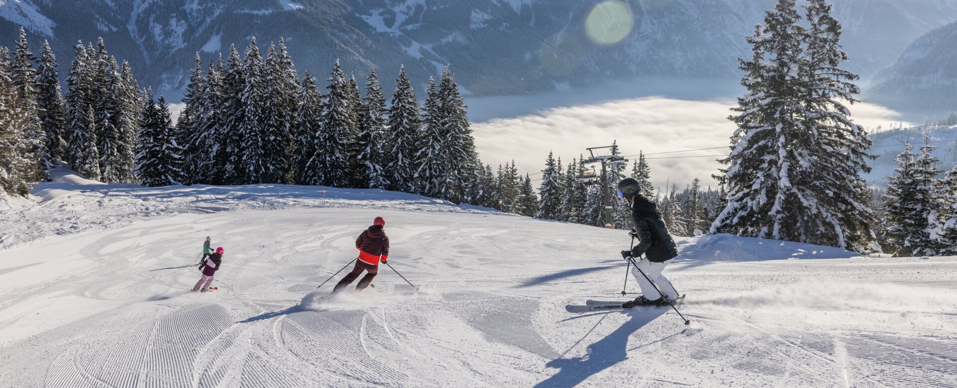 A family is skiing down the piste and the valley is covered in fog, while a bright blue sky can be seen above | © Michael Grössinger