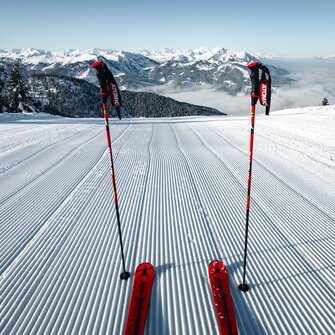 Two ski poles and red skis on freshly groomed ski slope with view of snow-covered alpine landscape | © Snow Space Salzburg Christian Schartner