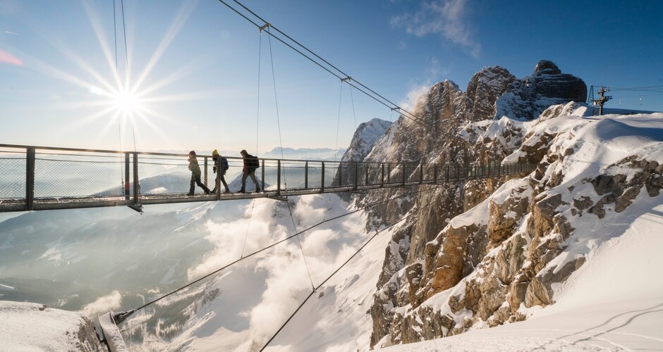 Four people cross a snowy suspension bridge at Dachstein in sunshine with panoramic valley view | © David McConaghy