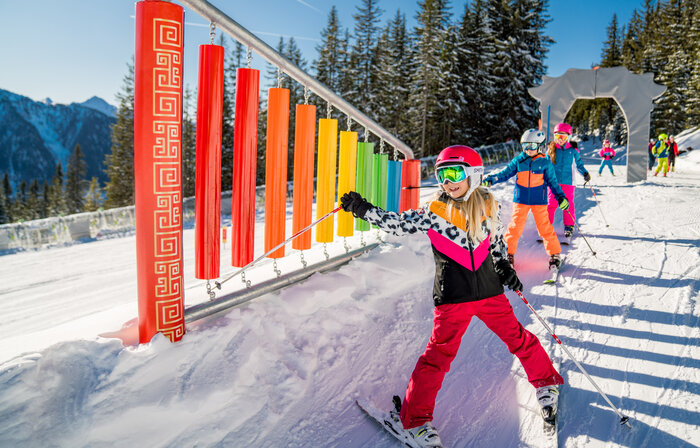 Children in colourful ski outfits pass xylophone in sunny kids’ area on Planai slope | © Christine Höflehner