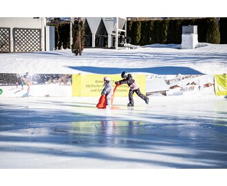 Two children in colourful gear and helmets ice skating with support aid on winter rink | © Gerhard Pilz